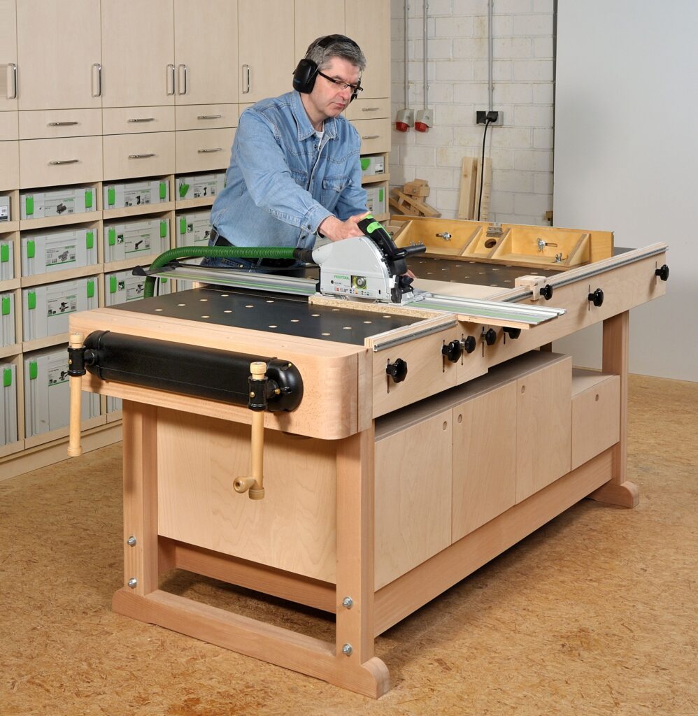 Tischlermeister Guido Henn steht mit Gehörschutz auf den Ohren an seiner selbstgebauten großen Multi-Werkbank und sägt mit der Handkreissäge ganz präzise ein Brett aus Spanplatte zu. 