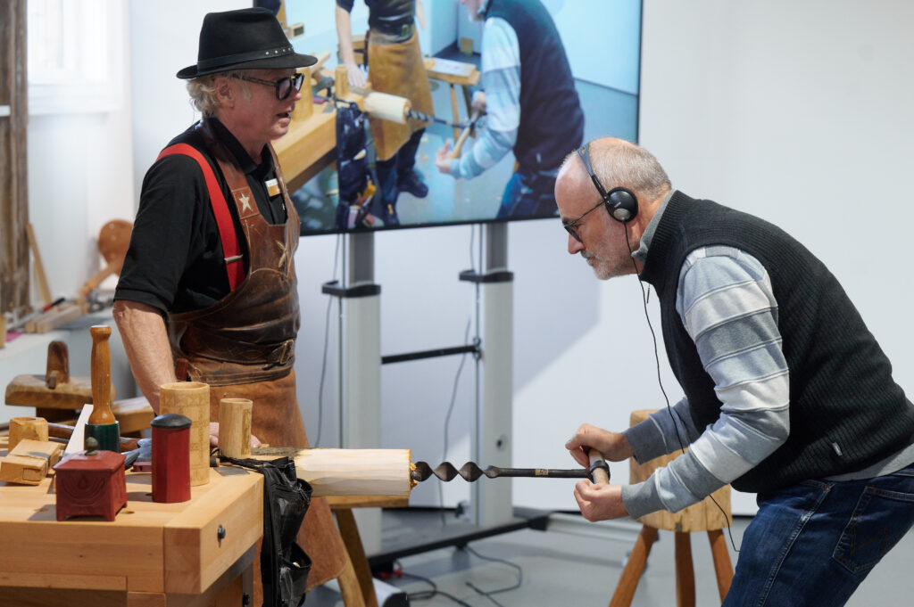 Ein Besucher des Kurses bei Jögge Sundqvist darf mit anpacken und dreht den großen Handbohrer für eine Dose ins Holz. Im Hintergrund steht der Schwede und erklärt.