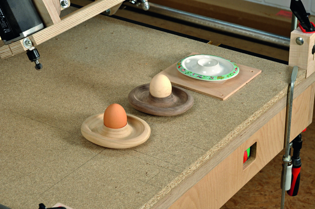 Fertige Ergebnisse des Kopierfräsens sind auf diesem Bild zu sehen. In einer Reihe stehen zwei fertig gefräste und geschliffene  Eierbecher aus Holz mit Eiern darin, dahinter die Kunststoff-Vorlage auf dem Werktisch, auf dem zuvor gefräst wurde.