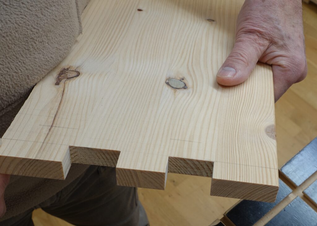 Eine Hand zeigt ein Brett mit einer Fingerzinkung in die Kamera. Diese Zinkung wurde sehr exakt mit der Stichsäge und einer passenden Vorrichtung hergestellt.
