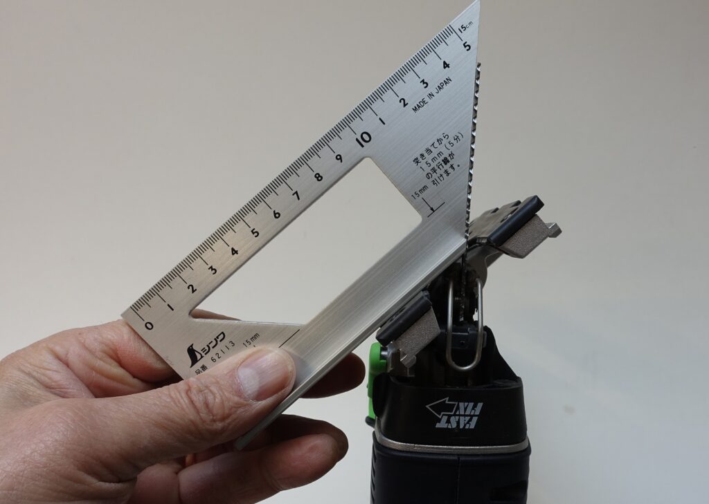 Eine Stichsäge steht auf dem Kopf. Der Tisch der Säge ist auf 45° eingestellt. Eine Hand hält eine Winkellehre zwischen Sägeblatt und Tisch, um zu zeigen, dass der Winkel exakt eingestellt ist. Weißer Hintergrund.