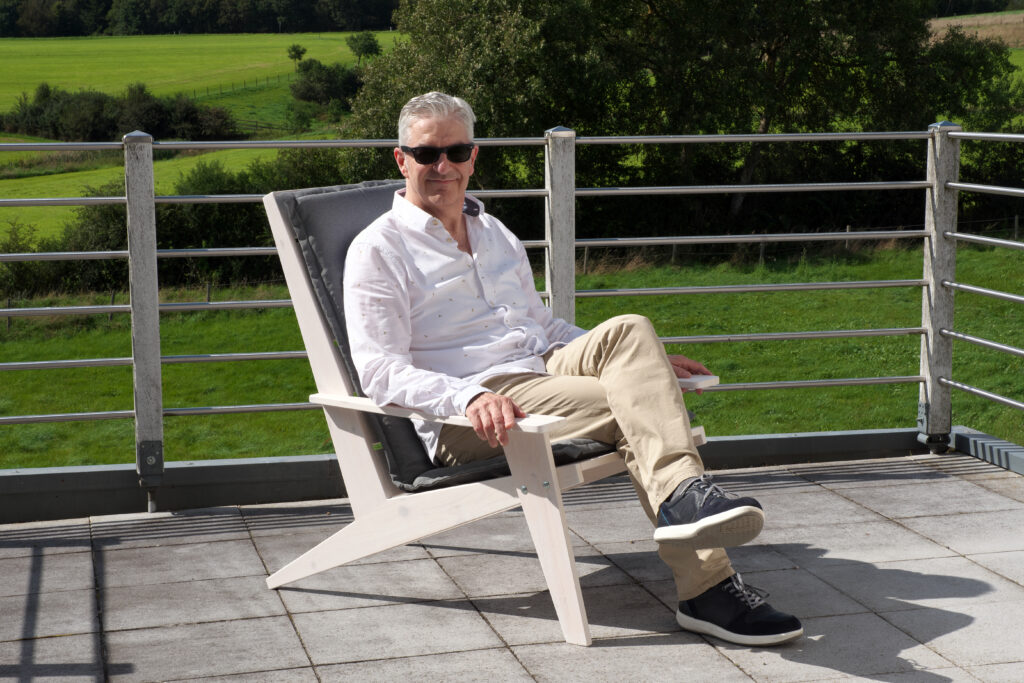 Autor Guido Henn sitzt auf dem von ihm gebauten Lounge-Sessel auf einer Terrasse in der Sonne. Er trägt helle sommerliche Kleidung und eine Sonnenbrille. Im Hintergrund hinter einer Terrassenbrüstung aus Edelstahl sind grüne Wiesen und Büschez u sehen.