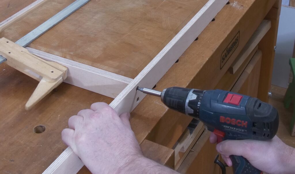 Eine Hand schraubt mit einem Akkuschrauber eine Schraube in eine selbstgebaute Vorrichtung zum beifräsen der Brett-Oberfläche.