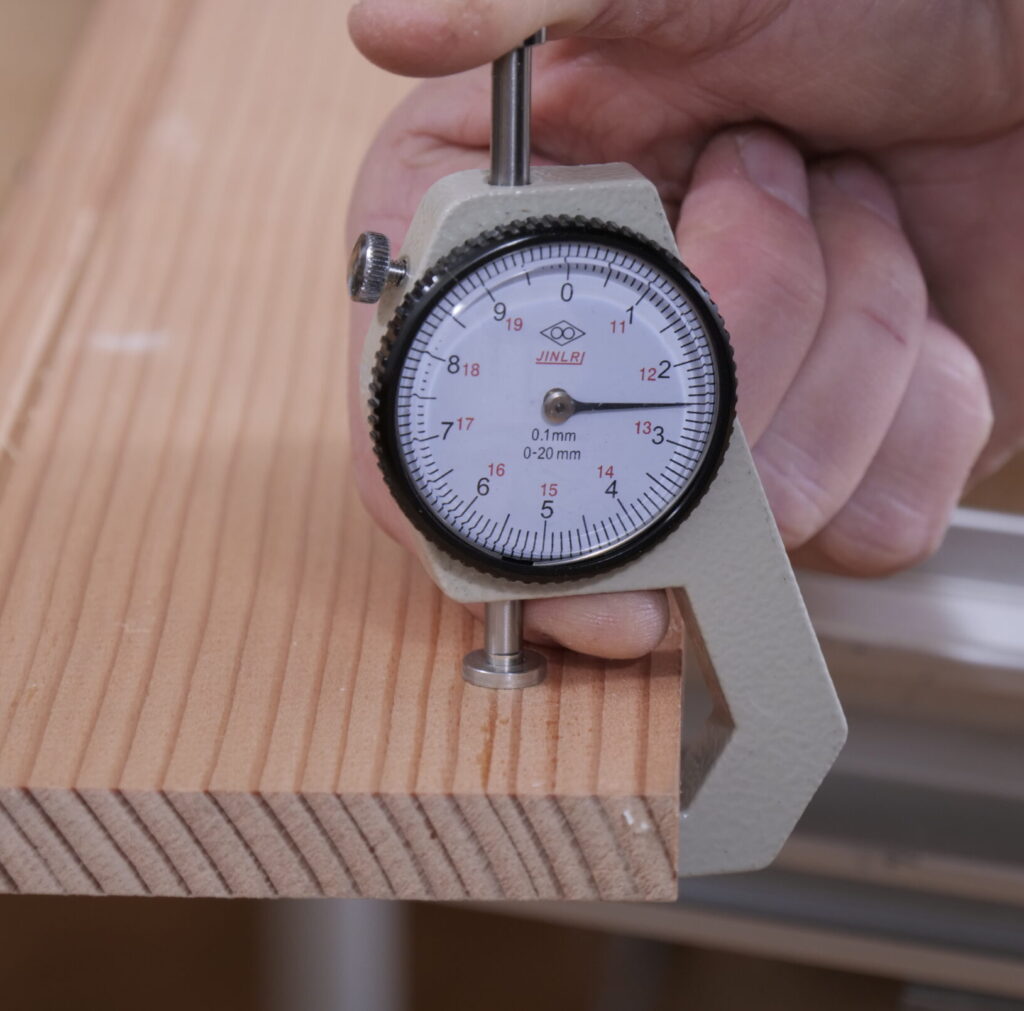 Das Bild zeigt eine Hand, die einen analogen Dickenmesser verwendet, um die Dicke eines Holzbretts zu messen. Der Dickenmesser trägt den Markennamen „JINRI“ und zeigt eine Skala von 0 bis 20 mm in 0,1-mm-Schritten. Das Holzbrett besteht aus hellem, naturfarbenem Holz mit sichtbarer Maserung und einer länglichen Nut auf der Oberfläche. Im Hintergrund sind unscharf eine Werkbank und Metallprofile erkennbar.