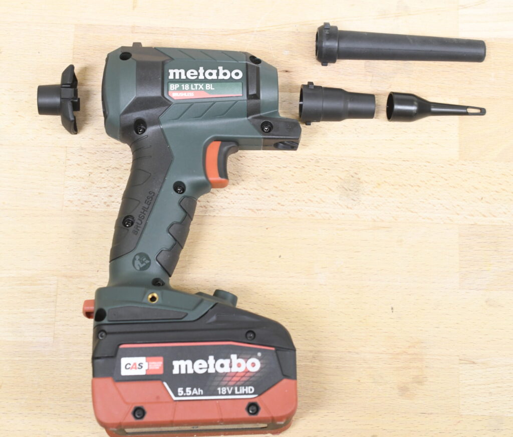 ine Metabo-Blaspistole mit rotem 5,5-Ah-18V-LiHD-Akku liegt auf einer Holzoberfläche. Daneben befinden sich drei verschiedene schwarze Düsenaufsätze, die für unterschiedliche Anwendungen ausgelegt sind.