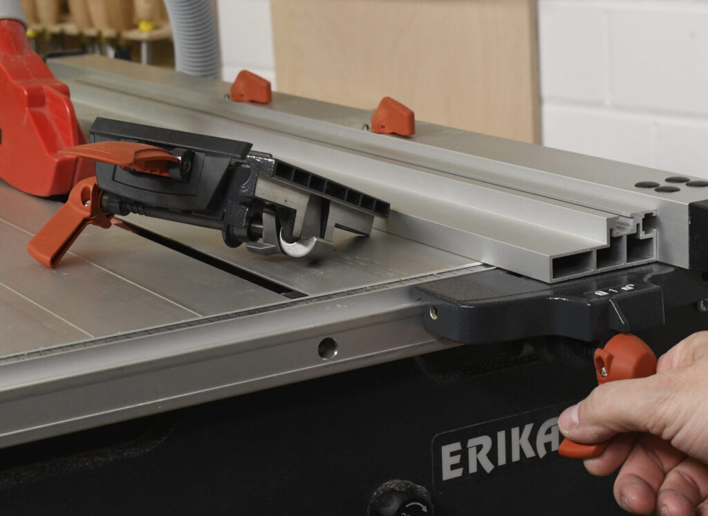 Das Bild zeigt eine Nahaufnahme des Klemmhebels für die Tischkreissäge der Marke Mafell, Modell „ERIKA“. Eine Hand greift einen roten Spannhebel an der Seite der Maschine, um eine Einstellung vorzunehmen. Auf der Sägeplatte befindet sich ein Parallelanschlag mit mehreren roten Bedienelementen zur Justierung.
