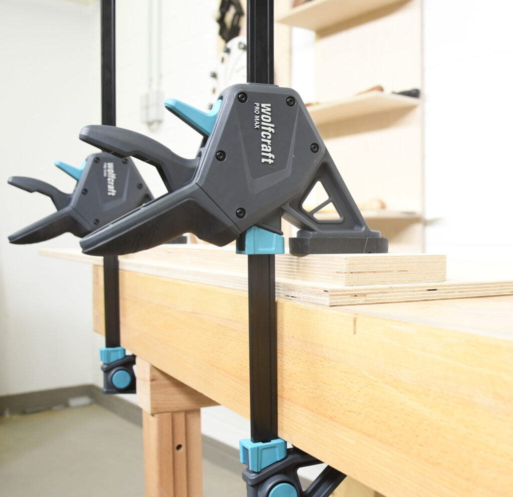 Das Bild zeigt zwei parallel an einer Werkbank befestigte Wolfcraft Pro Max Schraubzwingen. Die Schraubzwingen sind schwarz mit türkisfarbenen Akzenten und halten mehrere übereinandergelegte Sperrholzplatten fest. Die Werkbank besteht aus hellem Holz. Im Hintergrund sind Regale mit verschiedenen Werkzeugen und Materialien sichtbar.