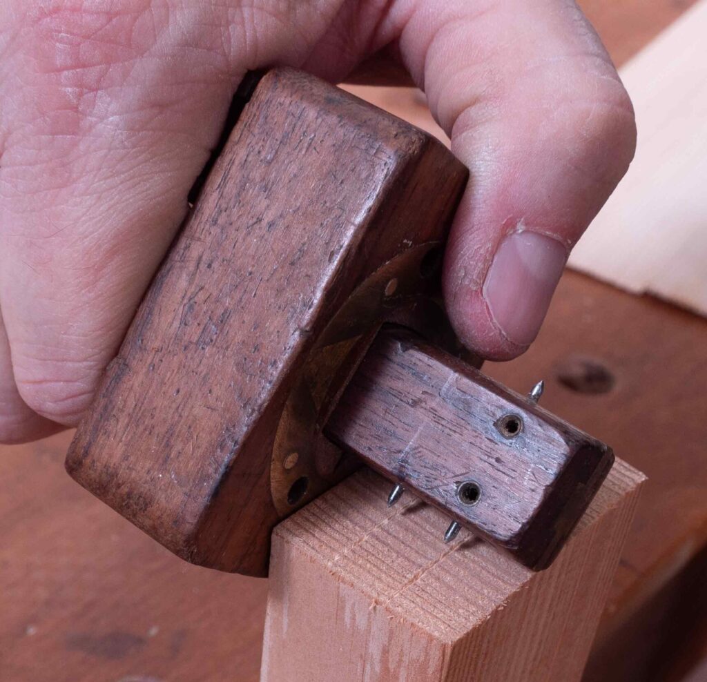 Eine Nahaufnahme einer Hand, die ein Holzwerkzeug, einen sogenannten Streichmaß, hält. Das Werkzeug besteht aus dunklem Holz und hat Metallstifte an der Unterseite, die feine Linien auf ein helles Holzstück ritzen. Die Hand wirkt kräftig und leicht staubig, vermutlich vom Holzstaub. Im Hintergrund ist eine Werkbank aus Holz sichtbar.