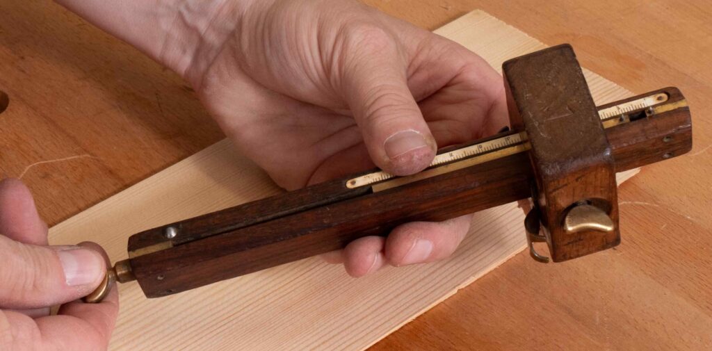 Zwei Hände halten ein hölzernes Streichmaß auf einem hellen Holzbrett. Eine Hand justiert das Messing-Einstellrad, während die andere das Werkzeug stabilisiert. Das Streichmaß hat eine Messskala aus Messing auf der Oberseite. Der Untergrund ist eine Holzwerkbank mit runden Löchern, die typisch für Werkbänke sind.
