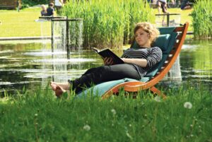 Eine Frau liegt auf einer Gartenliege an einem Teich und liest in einem Buch.