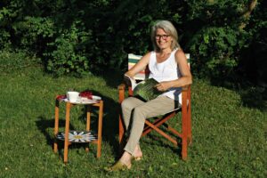Eine Frau sitzt auf einem Stuhl im Garten und hält ein Buch in den Händen. Daneben stehe ein Tablett-Tisch, auf dem eine Tasse steht.