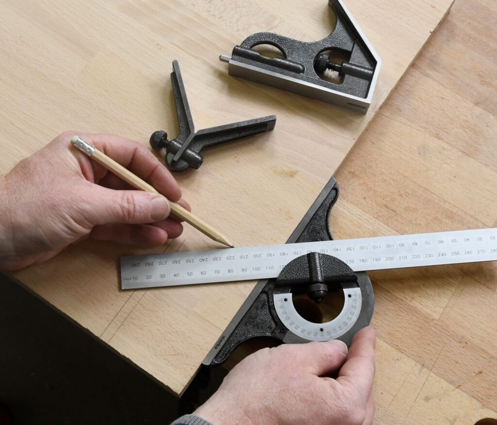 Eine Hand zeichnetn mit einem Bleistift und einem Kombinationswinkel eine Linie auf eine Holzplatte, die auf einer Werkbank liegt.