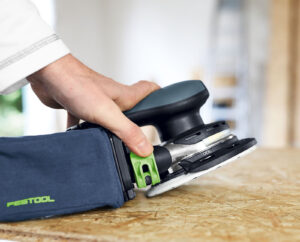 Eine Hand hält einen Akku-Exzenterschleifer mit Staubfangbox auf eine Arbeitsplatte.