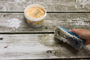 Eine Hand hält eine grobe Bürste, umd damit eine Holzoberfläche von Verschmutzungen zu befreien. Zu sehen ist außerdem eine geöffnete Dose mit Handwaschpaste.