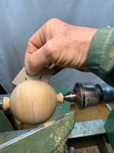 Eine Hand hält eine Schablone an eine Holzkugel, die in eine Drechselbank eingespannt ist.