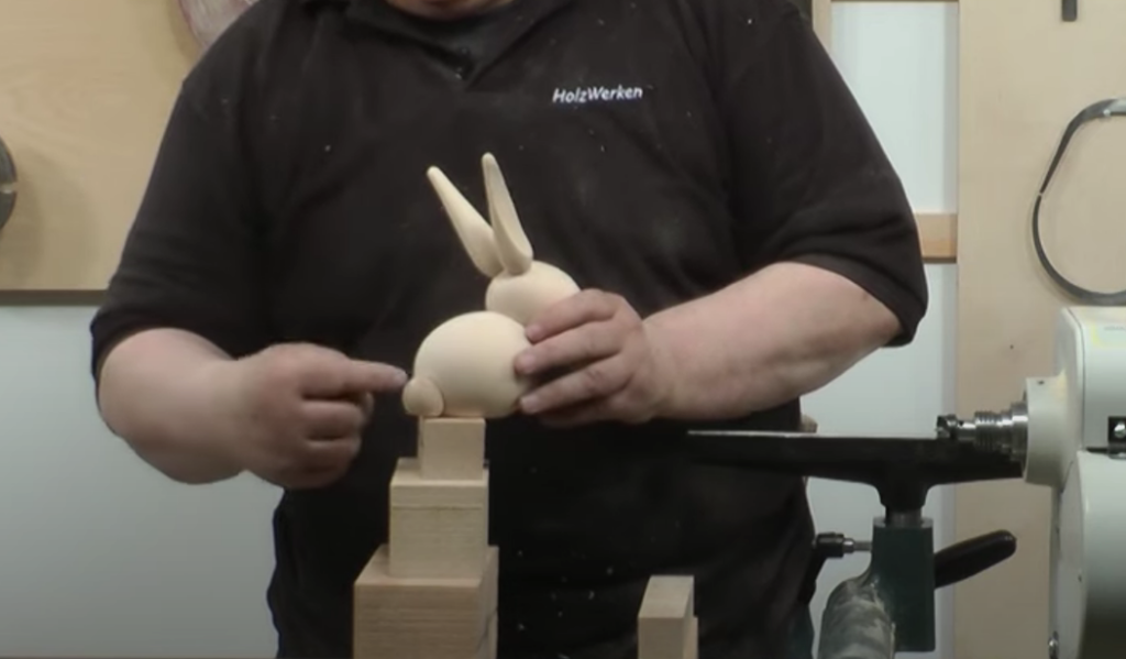 Ein Mann deutet mit dem Zeigefinger der rechten Hand auf eine fertige Osterhasenfigur. Darunter die Rohling in Klotzform.