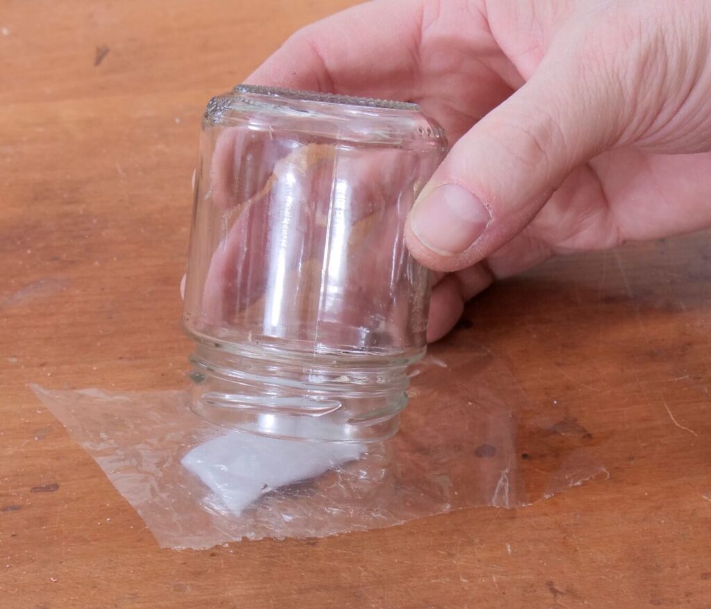 Eine Hand stülpt ein Glas über eine Kompresse mit Lösemittel, die unter einem Stück Zellophan liegt.