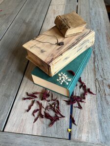 Drei gestapelte Bücher auf Holztisch, umgeben von roten getrockneten Blüten und kleinen weißen Blüten.
Zwei der Bücher sind aus Holz geschnitzt.