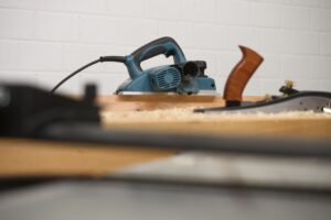 Im Hintergrund steht ein blauer Elektrohobel, davor (unscharf) ein Handhobel aus Metall.