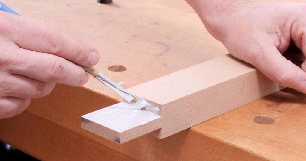 Ein Hand hält ein Werkstück aus Holz auf der Werkbank fest, während die andere Hand den Zapfen mit einem Pinsel und Leim bestreicht.