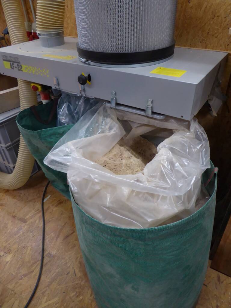 Absauganlage mit großem Filtersystem, darunter ein mit Sägespänen gefüllter Kunststoffsack in grünem Gartensack.