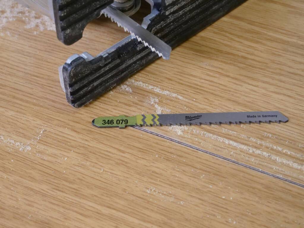 Stichsäge auf Holzplatte mit eingesetztem Sägeblatt, daneben ein zweites Milwaukee-Sägeblatt mit Aufschrift „Made in Germany“.