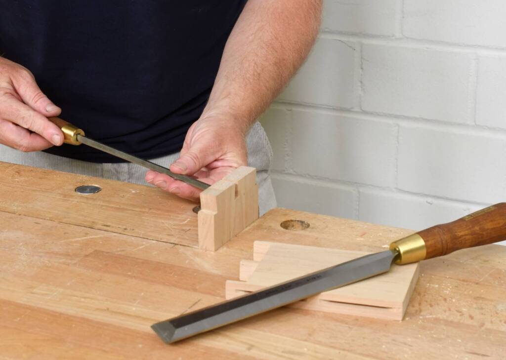 Person arbeitet mit einem Schälbeitel eine Fingerzinkenverbindung in einem Holzstück auf einer Werkbank ein.