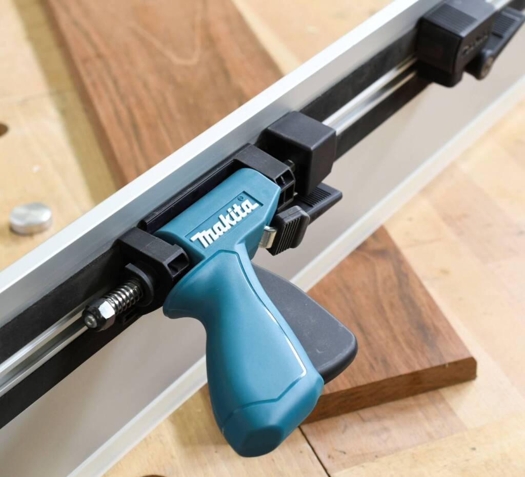 Makita-Führungsschiene mit Schnellspannzwinge fixiert ein Holzbrett auf einer Werkbank.