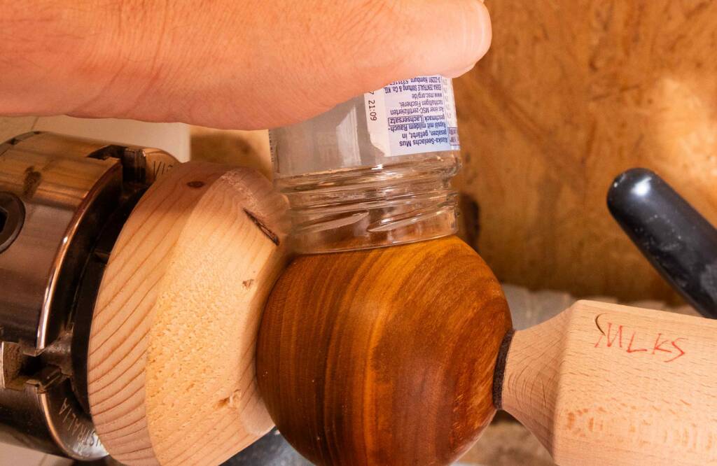 Hand hält ein Glas auf eine gedrechselte Holzkugel, die auf einer Drehbank eingespannt ist.