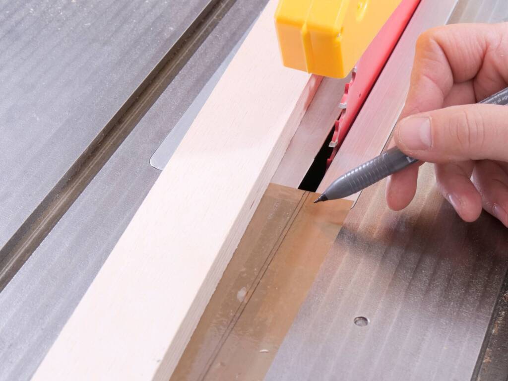 Hand markiert mit Stift eine Schnittlinie auf Holz vor Kreissägeblatt in Tischkreissäge.