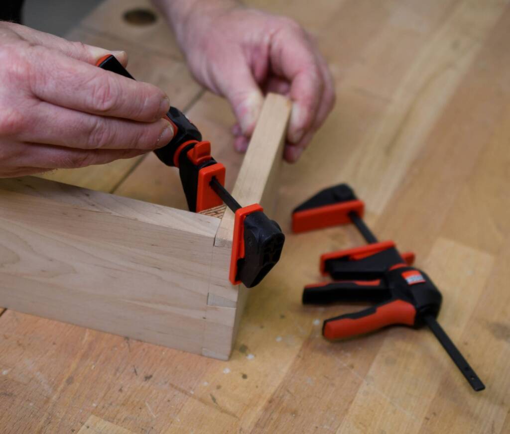 Holzverbindung mit Zinken wird mit kleiner Einhandzwinge auf Werkbank fixiert, zweite Zwinge liegt daneben.