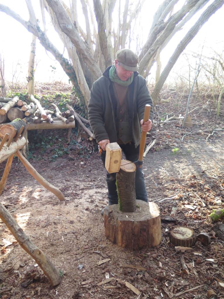 Mann spaltet Holz mit Keil und Hammer, umgeben von Baumstämmen und Werkbank