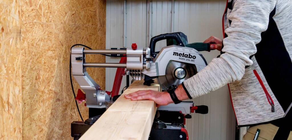 Mann mit Schutzbrille und Gehörschutz sägt langes Holzbrett an Kapp- und Gehrungssäge in Werkstatt.