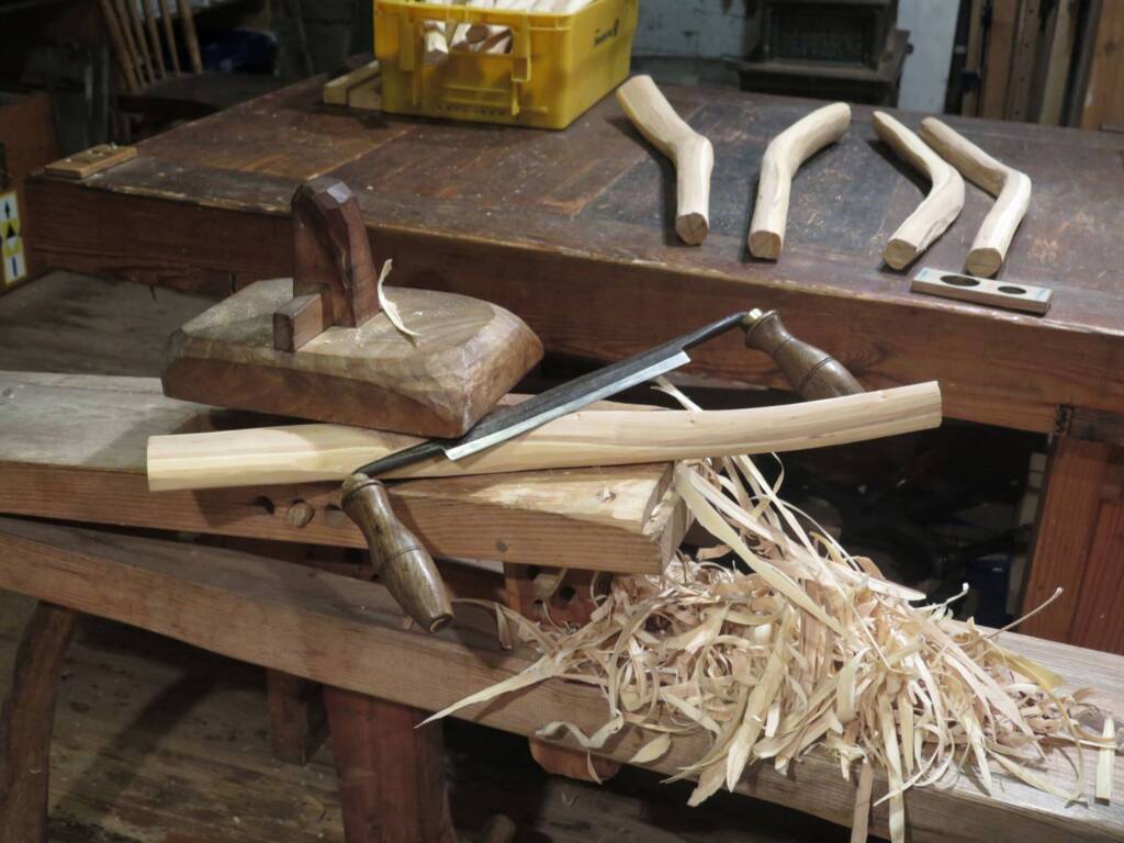 Auf einer Werkbank in einer Holzwerkstatt liegt ein eingespanntes Holzstück, das mit einem Handhobel bearbeitet wird. Holzspäne fallen am Rand der Werkbank herunter. Daneben liegen weitere Werkzeuge und zugeschnittene Holzrohlinge.