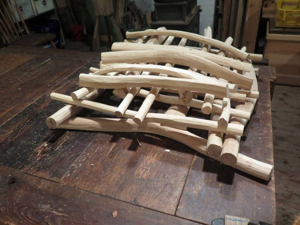 Mehrere gebogene und gerade, glatt bearbeitete Holzstäbe sind in einer losen Stapelstruktur auf einer Werkbank angeordnet. Die Holzteile wirken vorbereitet für den Bau eines größeren Werkstücks.
