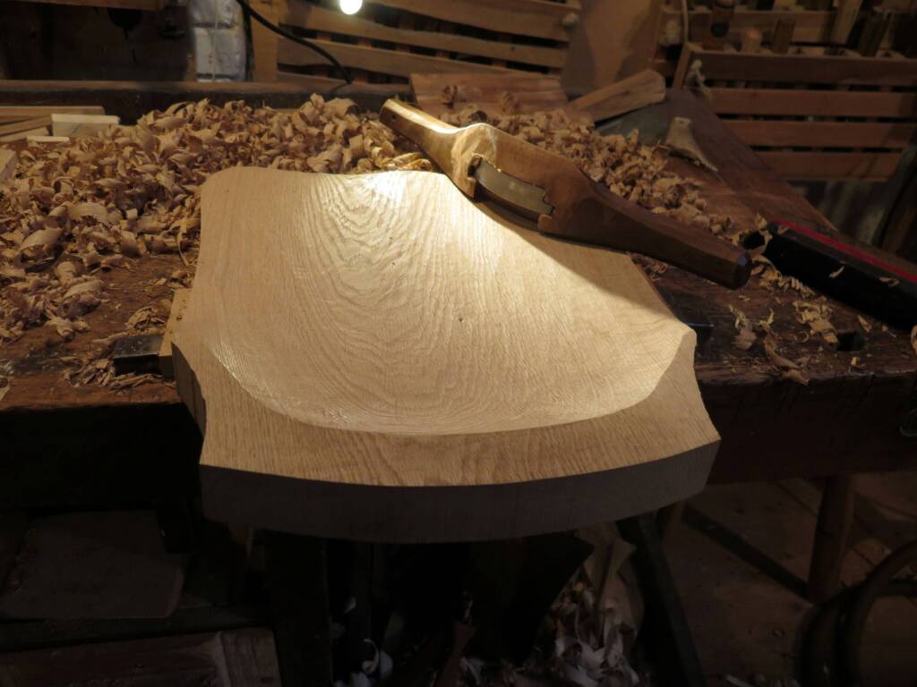 Ein ausgehöhltes Holzbrett liegt auf einer Werkbank und wurde mit einem Handhobel bearbeitet. Rundherum befinden sich zahlreiche Holzspäne, und ein Werkzeug liegt neben dem Werkstück.